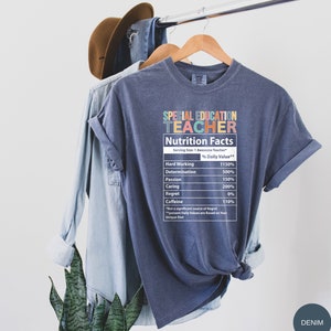 Op de afbeelding: Een blauw t-shirt met een afbeelding van een voedingsschema voor een "leraar voor speciaal onderwijs". Het schema geeft het percentage dagelijkse waarde voor hard werken, vastberadenheid, passie, zorg, spijt en cafe&iuml;ne.