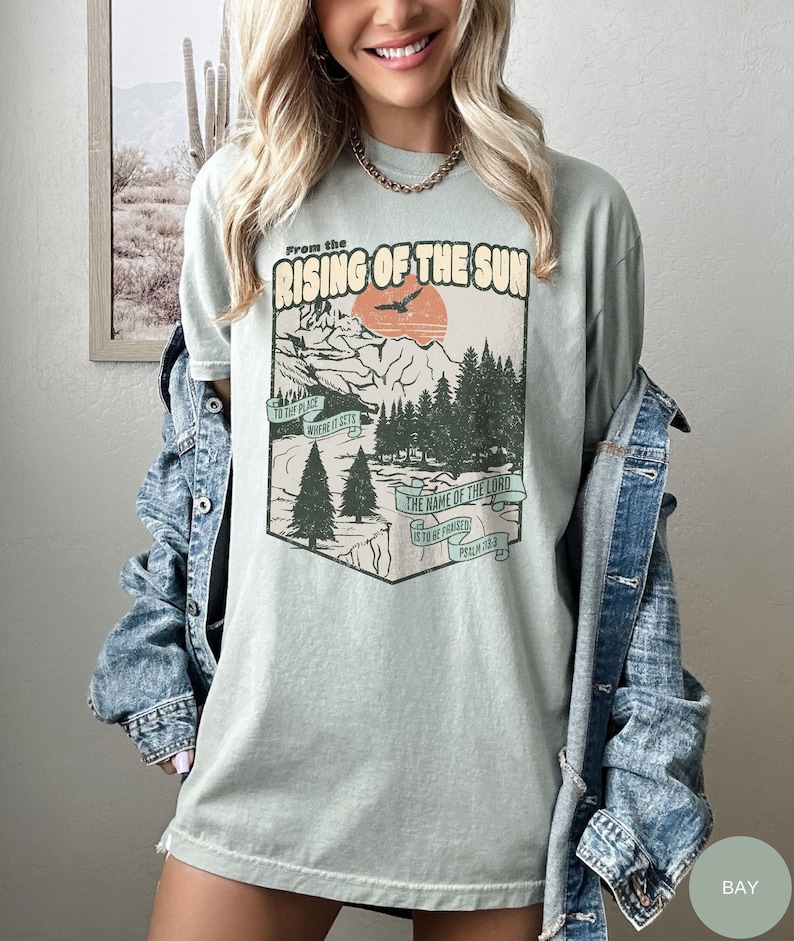 A model is wearing a bay colored shirt with a retro graphic that says from the rising of the sun to the place where it sets the name of the Lord is to be praised, psalm 113:3. Graphic shows mountains, river, trees, sunset, and a bird.