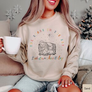 May include: A beige sweatshirt with a colorful graphic design featuring a nativity scene and the text "The Stars in the Sky Look down where He lay".