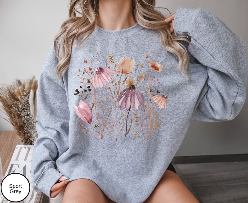 May include: A gray sweatshirt with a floral print featuring pink, brown, and white flowers. The sweatshirt has a crew neck and long sleeves.