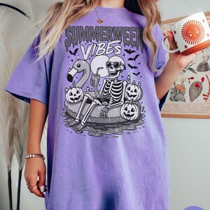 May include: Violet short-sleeved t-shirt with the text "Summerween Vibes" above a skeleton wearing sunglasses, relaxing on an inflatable ring with a flamingo and pumpkins. The skeleton is holding a drink. Bats are flying around the design.