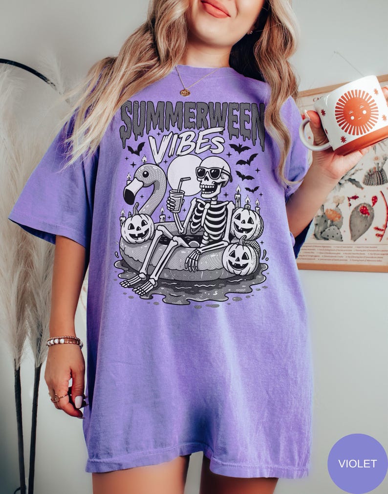 May include: Violet short-sleeved t-shirt with the text "Summerween Vibes" above a skeleton wearing sunglasses, relaxing on an inflatable ring with a flamingo and pumpkins. The skeleton is holding a drink. Bats are flying around the design.