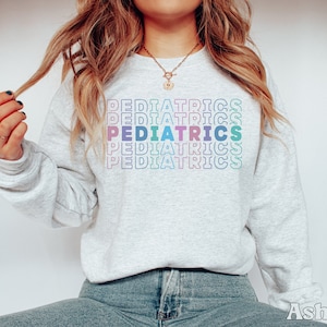 K&ouml;nnte beinhalten: Ein graues Sweatshirt mit dem Wort "PEDIATRICS", das in Regenbogenfarben wiederholt wird.