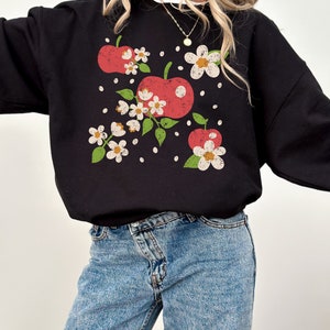A model wears a black sweatshirt with a retro inspired and slightly distressed graphic of 3 red apples surrounded by delicate cream flowers and some cream colored apple seeds around.