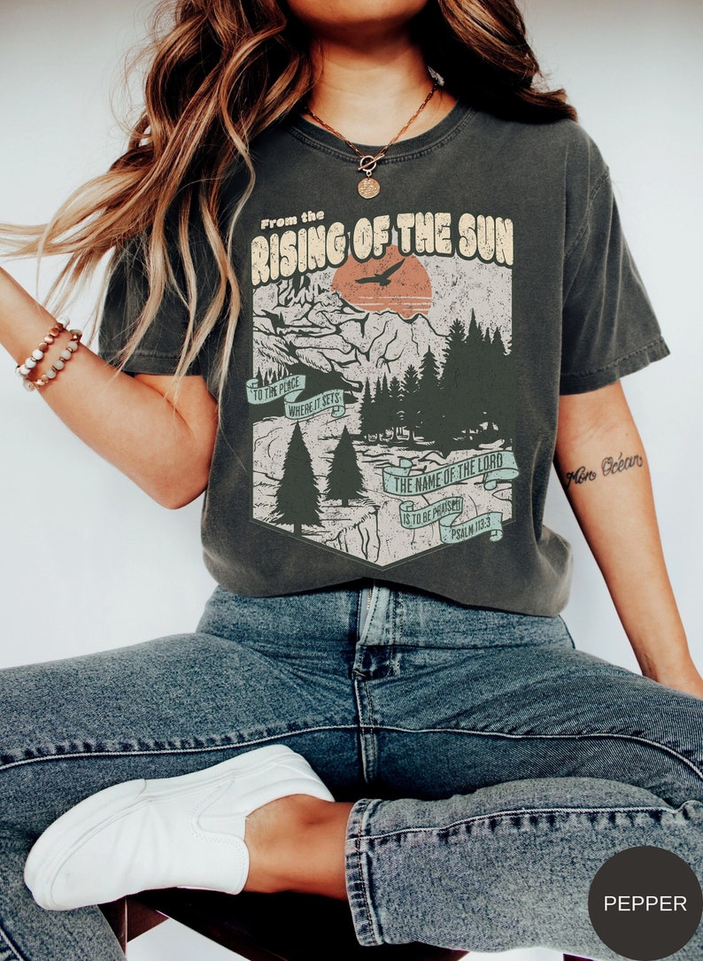 May include: A dark grey t-shirt with a vintage graphic design featuring a mountain landscape, a bird flying over a setting sun, and the text "From the Rising of the Sun to the Place Where It Sets The Name of the Lord is to be Praised Psalm 113:3".