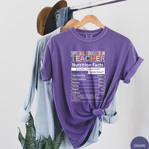 Op de afbeelding: Paars T-shirt met een afbeelding van een voedingsschema-etiket met de tekst "Special Education Teacher". Het etiket geeft de dagelijkse waarde van hard werken, vastberadenheid, passie, zorg, spijt en cafe&iuml;ne weer. De tekst "GRAPE" staat in de rechterbenedenhoek van de afbeelding.