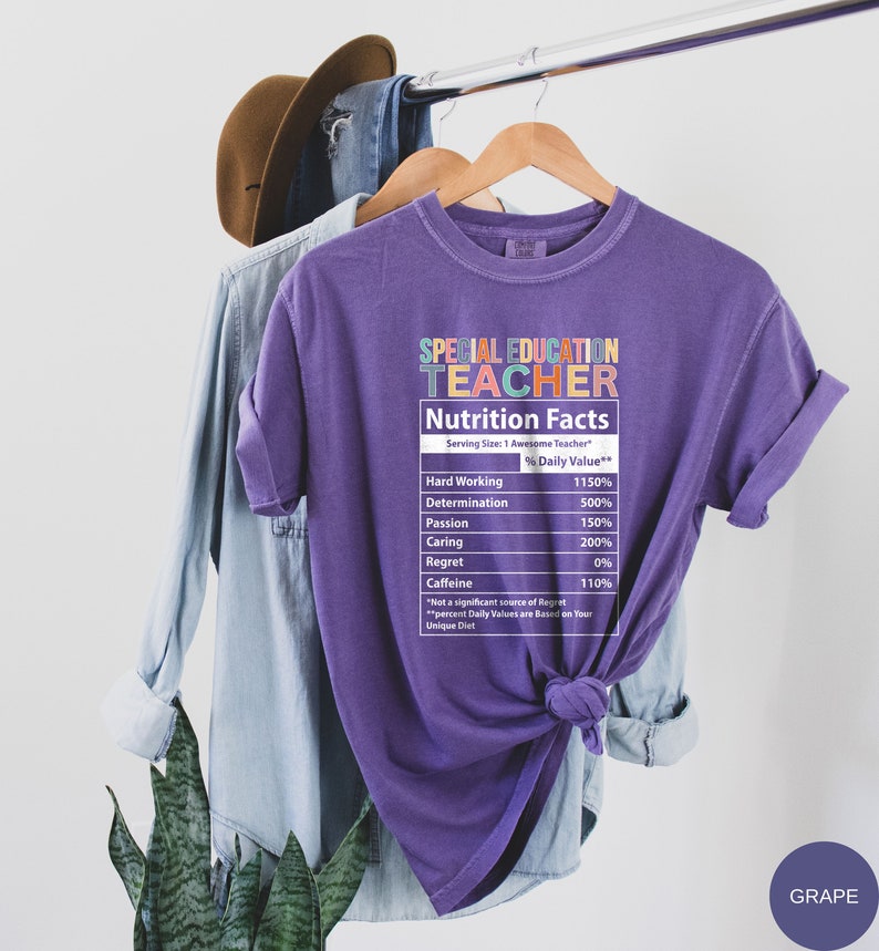 Op de afbeelding: Paars T-shirt met een afbeelding van een voedingsschema-etiket met de tekst "Special Education Teacher". Het etiket geeft de dagelijkse waarde van hard werken, vastberadenheid, passie, zorg, spijt en cafe&iuml;ne weer. De tekst "GRAPE" staat in de rechterbenedenhoek van de afbeelding.
