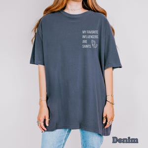 May include: A dark blue, oversized t-shirt featuring the white text "MY FAVORITE INFLUENCERS ARE SAINTS" on the upper back. The shirt has short sleeves and a loose fit. The word "Denim" is printed in a stylized font at the bottom.