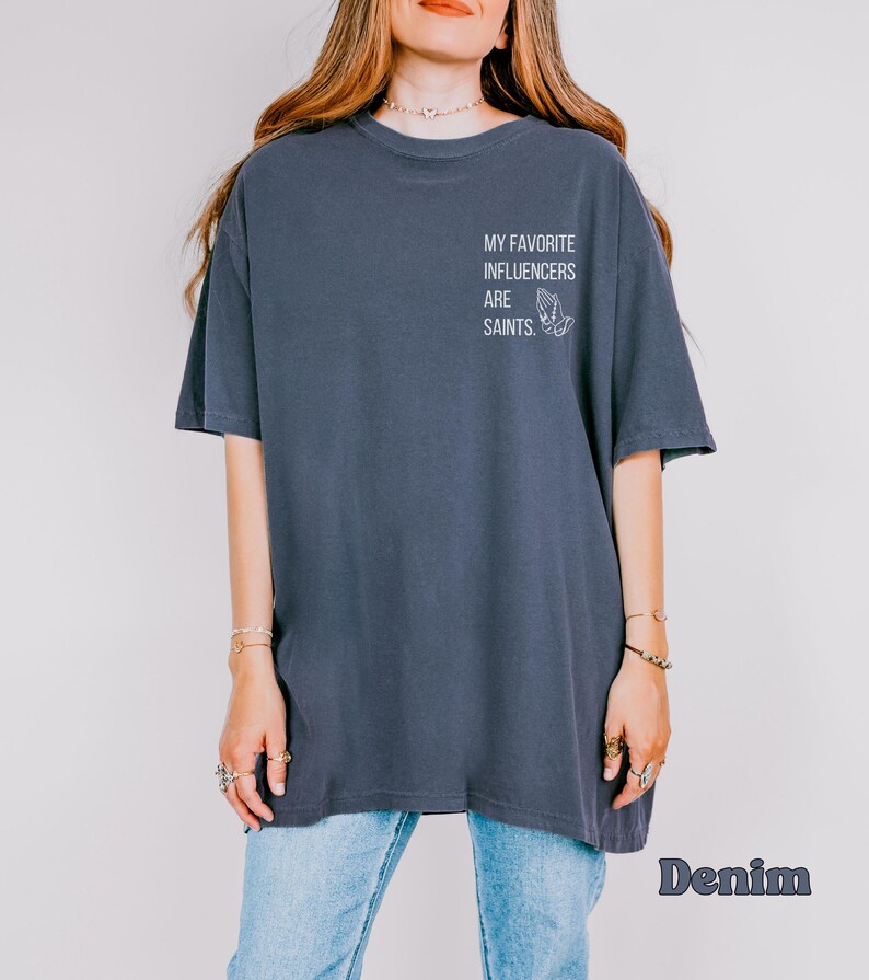 May include: A dark blue, oversized t-shirt featuring the white text "MY FAVORITE INFLUENCERS ARE SAINTS" on the upper back. The shirt has short sleeves and a loose fit. The word "Denim" is printed in a stylized font at the bottom.