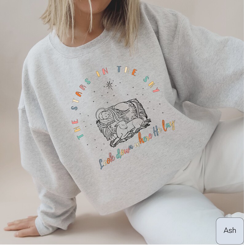 May include: A gray sweatshirt with a colorful graphic design featuring a nativity scene and the text "The stars in the sky look down where He lay".