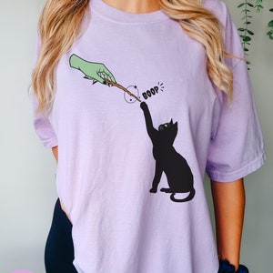 May include: A lavender t-shirt with a black cat reaching for a wand held by a green hand. The text "BOOP!" is above the cat.