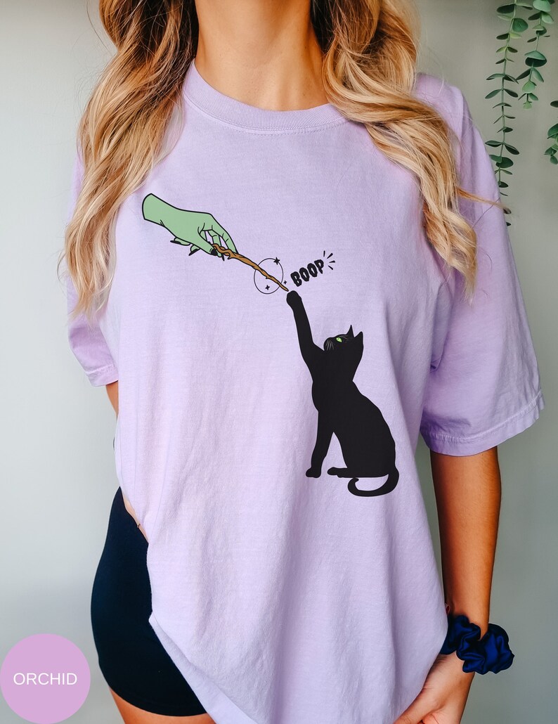 May include: A lavender t-shirt with a black cat reaching for a wand held by a green hand. The text "BOOP!" is above the cat.