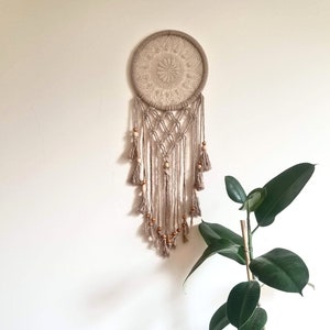 May include: A beige macrame dream catcher with wooden beads and tassels hanging from a circular base. The dream catcher is hanging on a white wall with a green plant in the foreground.