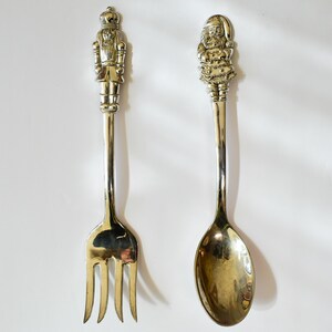 May include: Two decorative serving utensils. A fork with a nutcracker figure on the handle and a spoon with a Santa Claus figure on the handle. Both are made of a shiny, gold-colored metal.