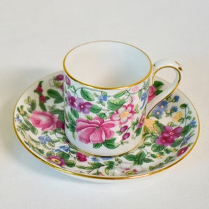 May include: A white porcelain teacup and saucer with a floral pattern in shades of pink, purple, blue, and green. The cup and saucer have a gold trim.