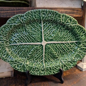May include: A green ceramic serving platter shaped like a cabbage leaf. The platter has detailed veining and a white accent at the center. It is displayed on a black stand, showcasing its unique design.