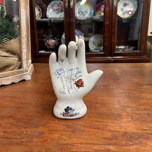 May include: A cream-colored ceramic palmistry hand figurine. The hand features black line drawings of palm lines, a red rose, and a diamond. A lightning bolt is on the index finger. The base of the hand has the text "The Palm".