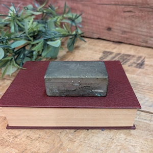 May include: A small, rectangular metal box with a tarnished, aged appearance. It sits atop a closed, burgundy-colored book with a textured cover. The background features a wooden surface and a sprig of green foliage.