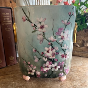 May include: A pale blue ceramic vase with a floral design of pink and white blossoms. The vase has gold accents on the handles and pink feet.