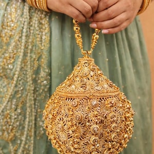 May include: Ornate gold-colored handbag with intricate floral and geometric patterns. The bag has a rounded shape, a tassel, and a chain handle. Gold bangles adorn the wrists. The background is a soft green fabric.