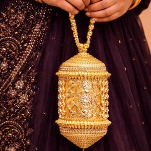 Op de afbeelding: Een sierlijke goudkleurige handtas met ingewikkelde details en sprankelende versieringen. De tas heeft een kralen hengsel en wordt tegen een donkerpaarse stoffen achtergrond gehouden. Gouden armbanden sieren de polsen.