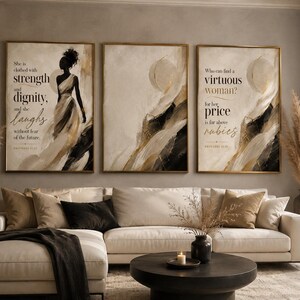 Op de afbeelding: Een set van drie ingelijste wandkunstprints met abstracte penseelstreken in zwart, goud en crème. De linkerprint toont een silhouet van een persoon in een vloeiende jurk met de tekst "She is clothed with strength and dignity, and she laughs without fear of the future." De rechterprint bevat de tekst "Who can find a virtuous woman? for her price is far above rubies."