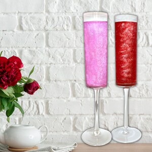 May include: Two champagne flutes filled with pink and red glitter liquid. The flutes are on a white brick background.