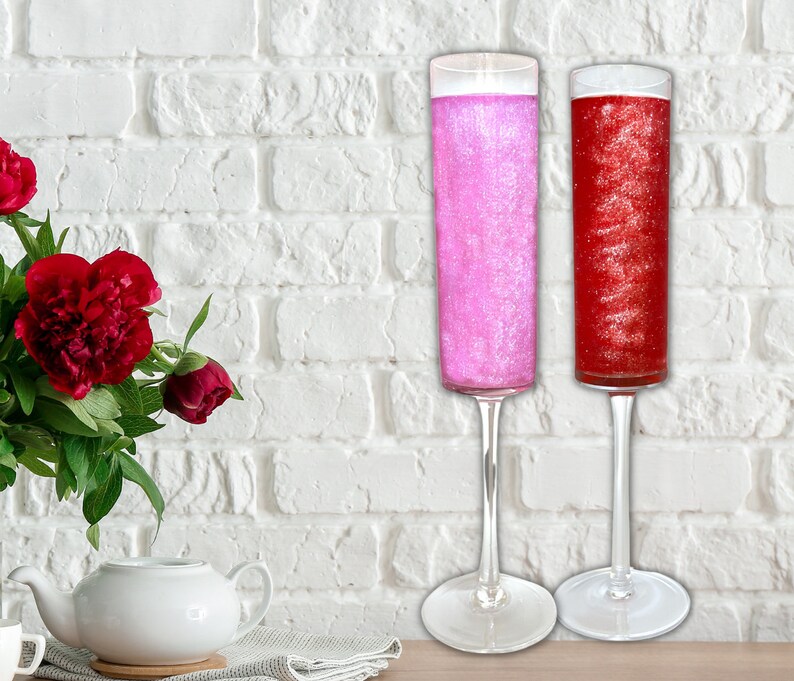 May include: Two champagne flutes filled with pink and red glitter liquid. The flutes are on a white brick background.