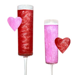 May include: Two tall clear glass champagne flutes filled with pink and red glitter liquid. A pink heart is next to the red glitter flute and a red heart is next to the pink glitter flute.