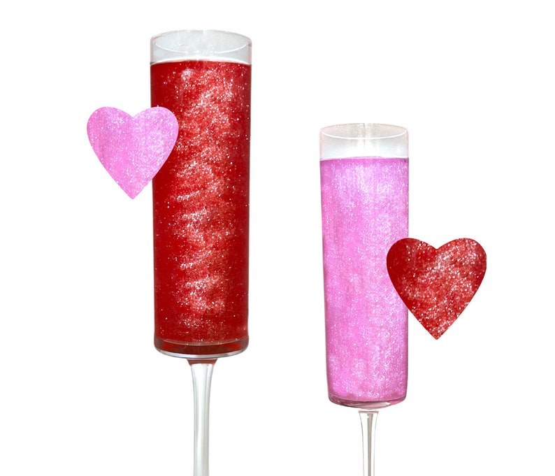 May include: Two tall clear glass champagne flutes filled with pink and red glitter liquid. A pink heart is next to the red glitter flute and a red heart is next to the pink glitter flute.