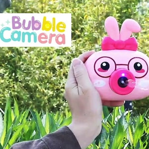 May include: Pink plastic bubble blower shaped like a bunny with a bow and glasses. The bubble blower has a red button to blow bubbles. The text "Bubble Camera" is in the top left corner of the image.