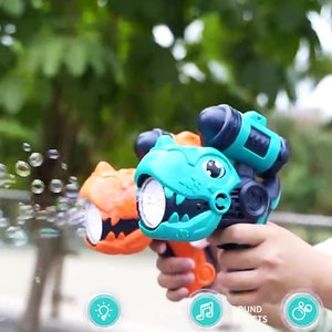 May include: Two green and orange dinosaur-shaped bubble blowers with lights and sound effects. The blowers are held by a person's hands and are blowing bubbles.