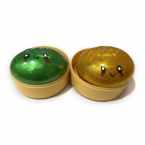 May include: Two glittery slime toys, one green and one gold, each in a tan container. The slime has a smiling face with rosy cheeks. The toys are on a white background.