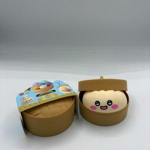 May include: Two brown plastic containers, one open and one closed, resembling bamboo steamers. The open container reveals a smiling, cartoon-style dumpling with pink cheeks. The closed container has a cardboard label with images of dumplings.