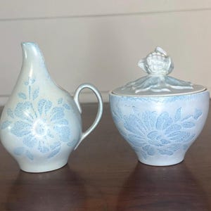 May include: A light blue and white ceramic creamer and sugar bowl set. The creamer has a handle and a spout. The sugar bowl has a lid with a decorative top. Both pieces feature a floral design.
