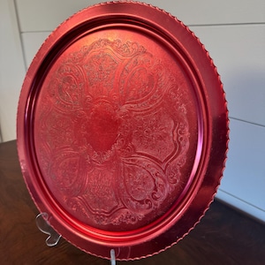 May include: A round, red metal tray with an ornate, embossed floral design. The tray has a scalloped edge and is displayed on a clear stand. The tray is a vibrant shade of red.