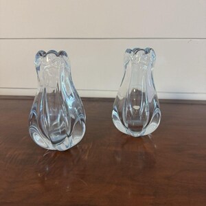 Könnte beinhalten: Zwei klare, hellblaue Glas-Blumenvasen mit gerüschtem oberem Rand. Die Vasen haben eine tulpenartige Form und stehen auf einer dunklen Holzoberfläche. Das Glas hat eine gerippte Textur, die das schlichte Design aufwertet.