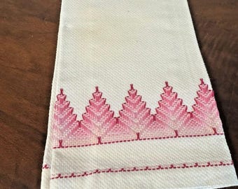 Puede incluir: Toalla blanca doblada con un borde bordado en rosa que presenta un diseño repetitivo en forma de árbol. El bordado es de un tono rosa más oscuro, creando un contraste con la tela blanca. La toalla está doblada cuidadosamente.