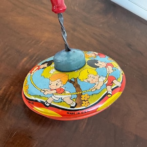May include: Vintage metal toy top with a red handle and a blue base. The top features a colorful illustration of children playing with a rope. The words "MADE IN U.S.A." are printed on the side.