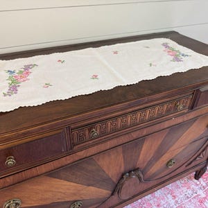 May include: A vintage wooden dresser with a decorative floral embroidered table runner. The dresser is dark brown with brass hardware and a sunburst design on the drawer fronts. The runner is white with colorful floral embroidery.