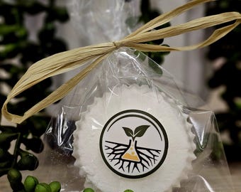 Könnte beinhalten: Ein weißes, rundes Produkt mit gewelltem Rand ist in durchsichtigem Zellophan verpackt und mit einer Raffiabandschleife gebunden. Das Produkt hat ein kreisförmiges Logo mit einem Pflanzen- und Wurzeldesign. Grünes Laub befindet sich im Vorder- und Hintergrund.