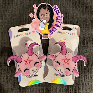 May include: Two pink glitter phone grips with a cartoon goat head design. Each grip has a red pentagram on the forehead and a white background. The text "PopGrip" is printed on the bottom of each grip.