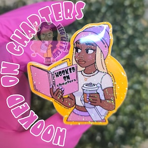 May include: A glittery, yellow and pink phone grip featuring a cartoon woman reading a pink book titled "Hooked on Chapters." The grip also includes the text "No Chapters" and a small illustration of a person holding a phone.