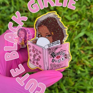 May include: A pink and yellow sticker with the words "BLACK CUTIE GIRLIE" in a bubble font. The sticker features a Black woman reading a pink book with lipstick marks and the words "Burn Book". The text "Dear Black girl, you are enough" is also visible.