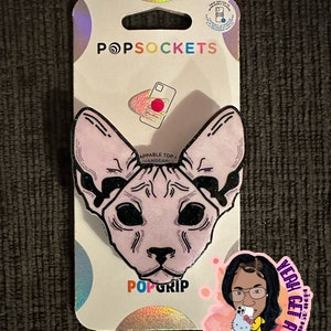 May include: A pink and black PopGrip phone accessory featuring a Sphynx cat design. The PopGrip is on a white card with the words "PopSockets" and "PopGrip" printed on it. The card also has a small image of a phone with a red circle on the screen.