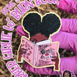 May include: A sticker featuring a cartoon illustration of a Black woman reading a pink book with the words "Burn Book" on the cover. The sticker has glitter accents and the text "Girlie W Darked Cute Swap None" around the edges.