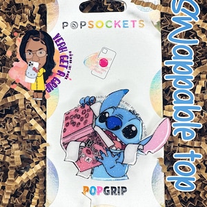 May include: A white phone grip with a blue and pink Stitch character holding a pink book with the text "Book Club" on it. The text "Swappable Top" is written in blue on the right side of the image. The text "PopGrip" is written in rainbow colors at the bottom of the image.