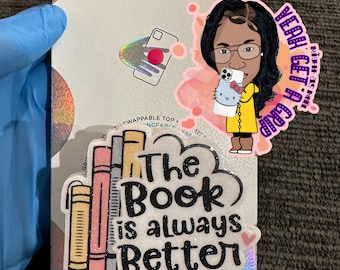 Op de afbeelding: Een witte PopSockets-telefoonhouder met een glinsterende sticker waarop staat "The Book is always Better" met een stapel boeken en een roze hart. De sticker bevindt zich op een witte achtergrond met een holografisch regenboog-effect.