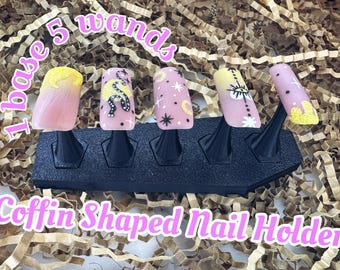 May include: A black coffin-shaped nail holder with five nail art designs. The nails feature pink, yellow, and white colors with various designs, including a snake, stars, and an eye. The text on the image says "1 base 5 wands" and "Coffin Shaped Nail Holder."