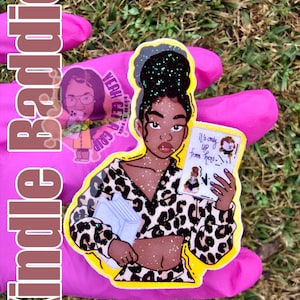 May include: A sticker featuring a Black woman with a high bun, wearing a leopard print outfit, holding a book and a sign that reads "It's only up from here." The sticker has glitter accents and a yellow border. The word "Kindle Baddie" is written vertically on the left.